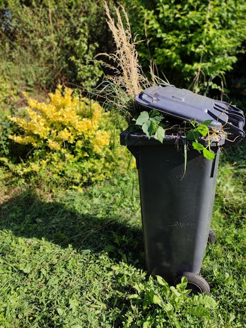 A black wheeled garden waste bin positioned on a grassy area, filled with freshly cut garden prunings, dried ornamental grasses, and green leafy stems. The background features dense, lush green bushes