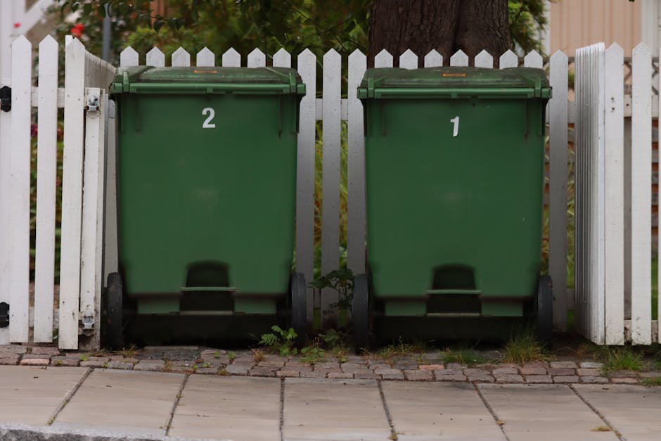 A black wheeled garden waste bin positioned on a grassy area, filled with freshly cut garden prunings, dried ornamental grasses, and green leafy stems. The background features dense, lush green bushes